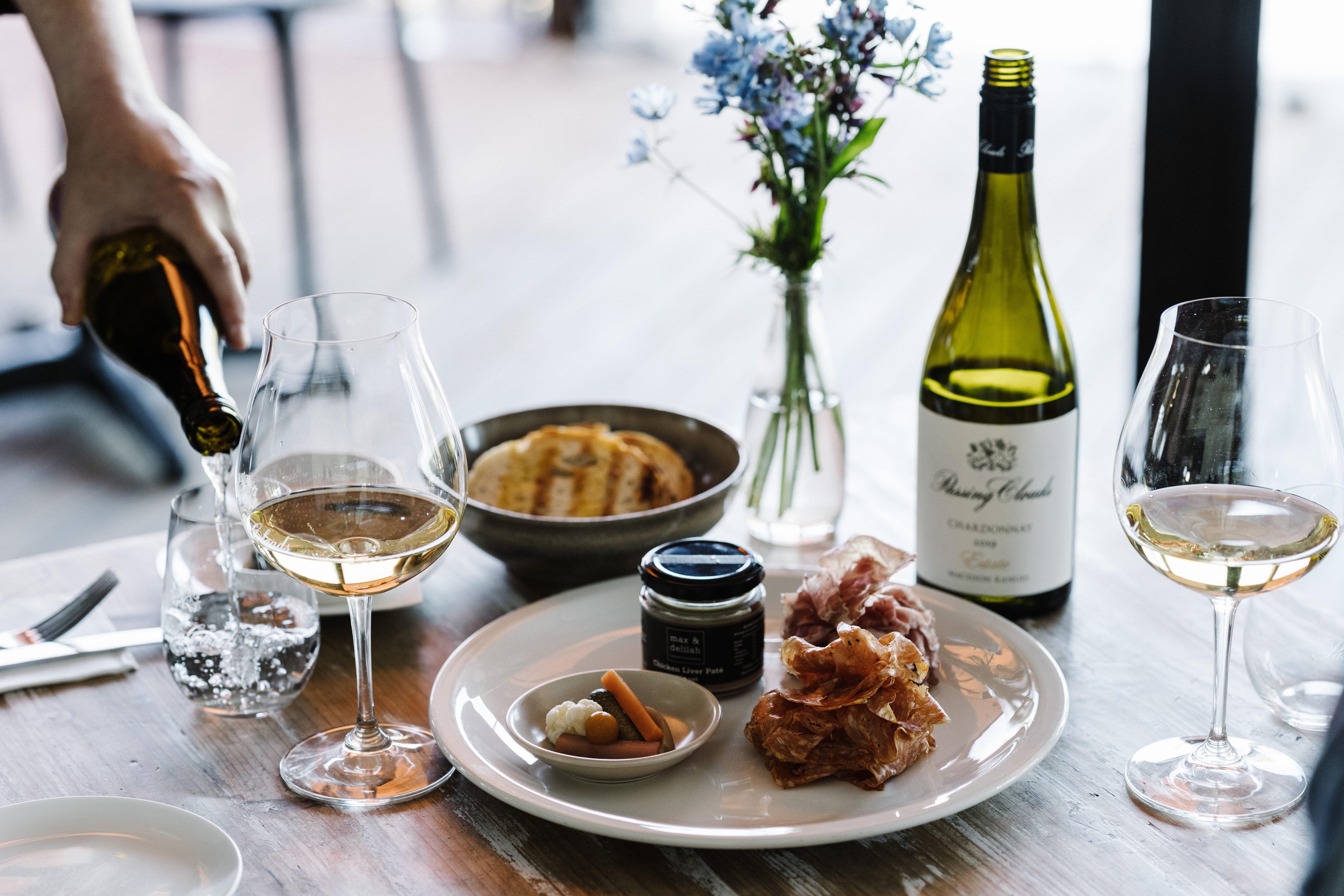 Table with wine glasses and food and a bottle of wine also a hand pouring wine